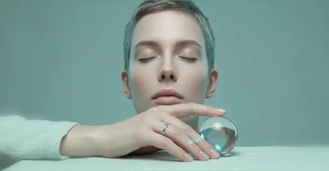 A woman with short gray hair and closed eyes resting her chin near a clear glass orb on a teal surface.