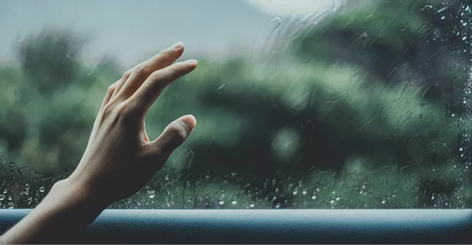 A hand reaching toward a rain-covered window with blurred green trees visible outside on a rainy day.