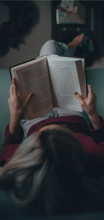 A woman lying on a couch reading an open book held above her face wearing a red sweater in a cozy indoor setting.