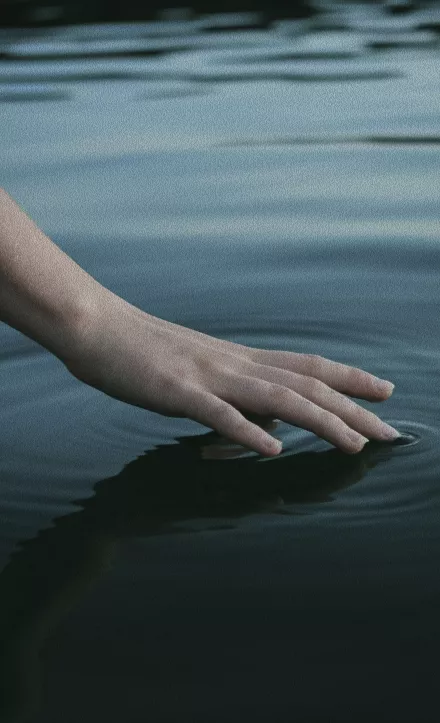 A hand gently touching the calm dark surface of still water creating small ripples in soft evening light.