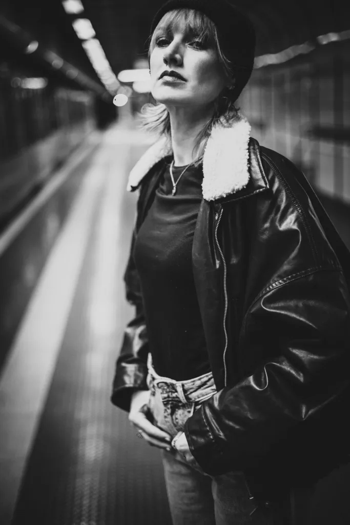 Frau mit Lederjacke und Baskenmütze steht auf einem U-Bahnsteig, Schwarz-Weiß-Porträt