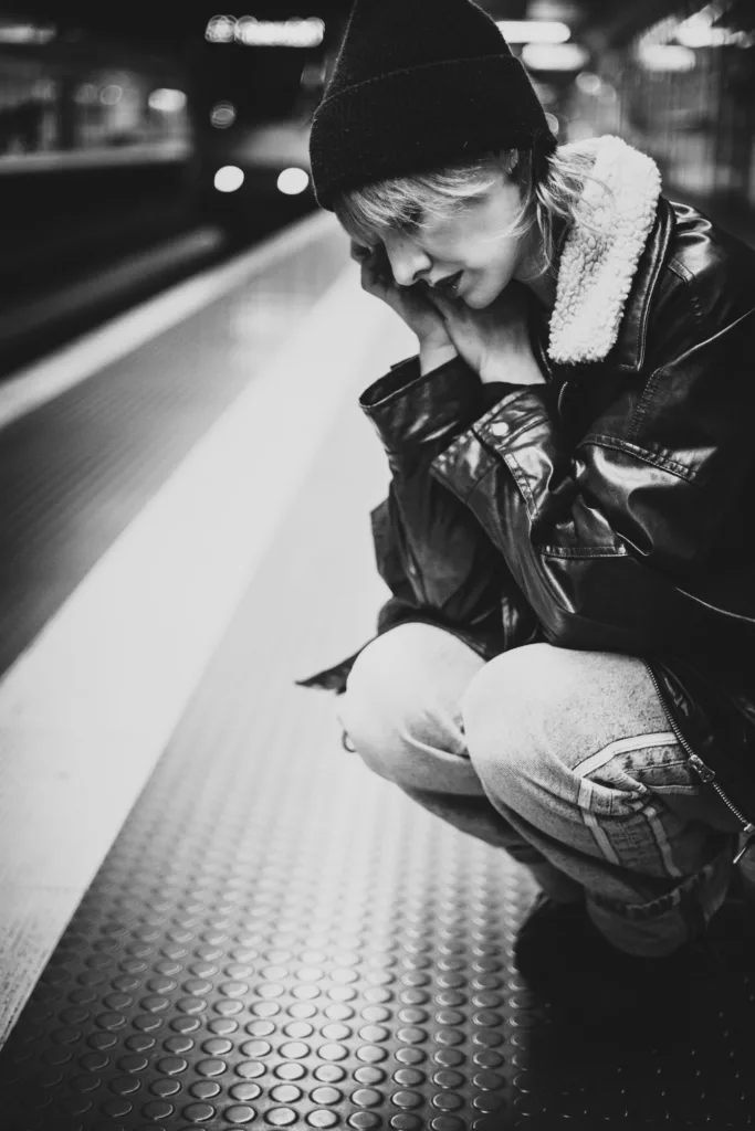Junge Frau in Lederjacke und Mütze hockt nachdenklich auf einem U-Bahnsteig