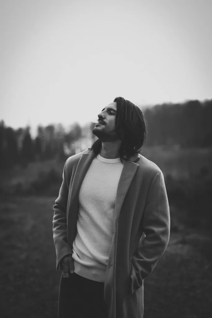 Mann mit langen Haaren und Mantel steht mit geschlossenen Augen in einer Landschaft, Schwarz-Weiß-Porträt